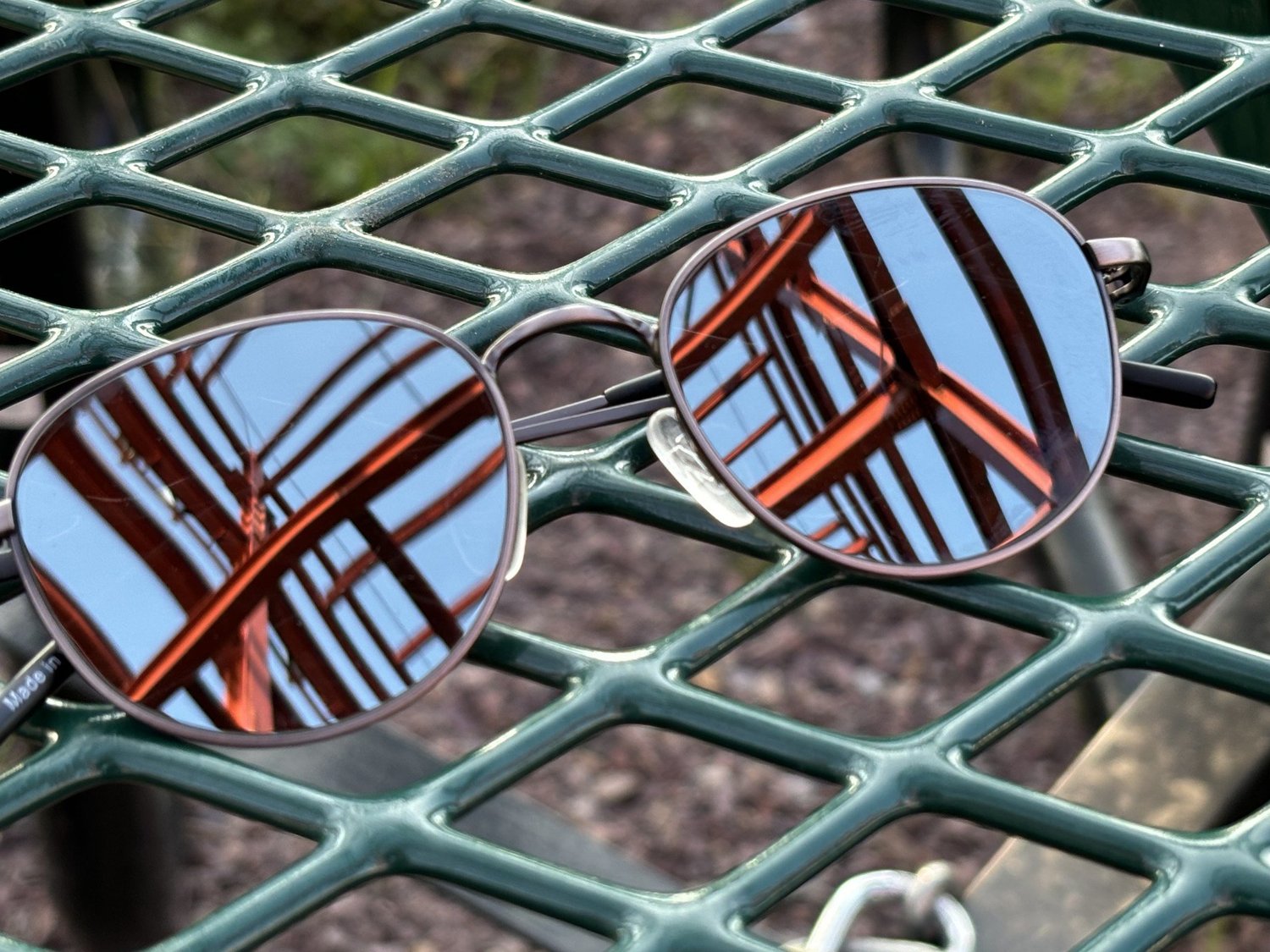 A pair of round sunglasses resting on a green metal mesh table, a red bridge reflected in the lenses. Photograph by Grant Williams.